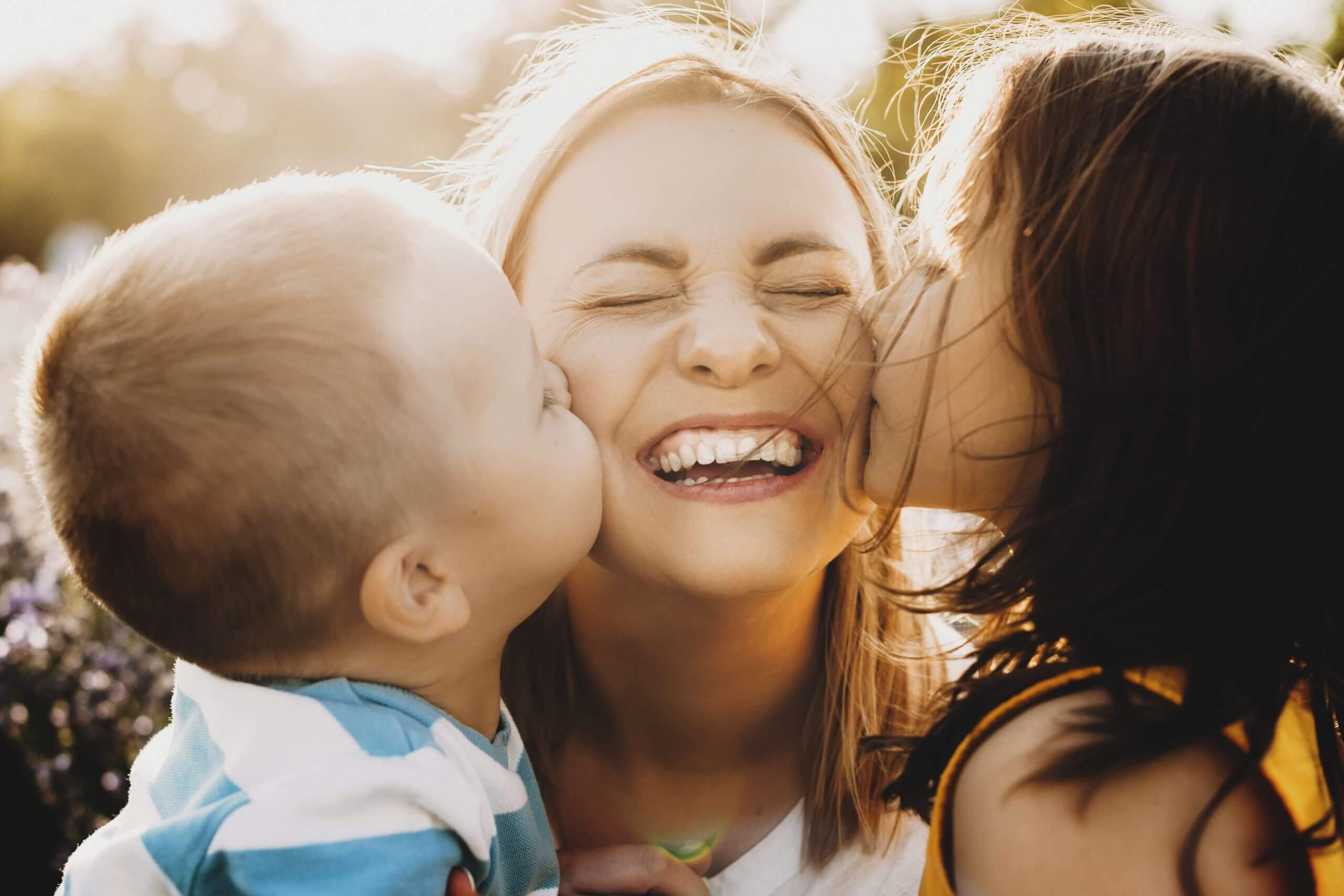 Imagem de duas crianças beijando as bochecas de uma mulher.